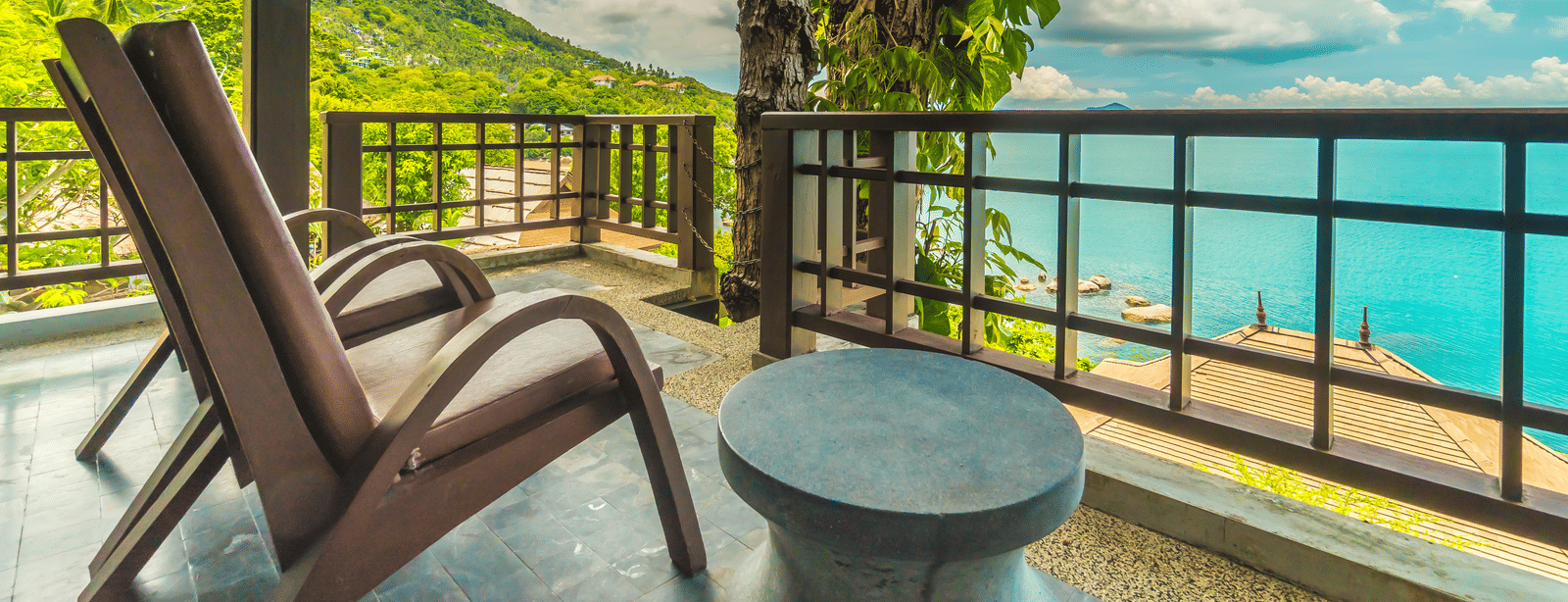 A balcony with 2 wooden lounge chairs, a small round table and a view of the sea and surrounding greenery.