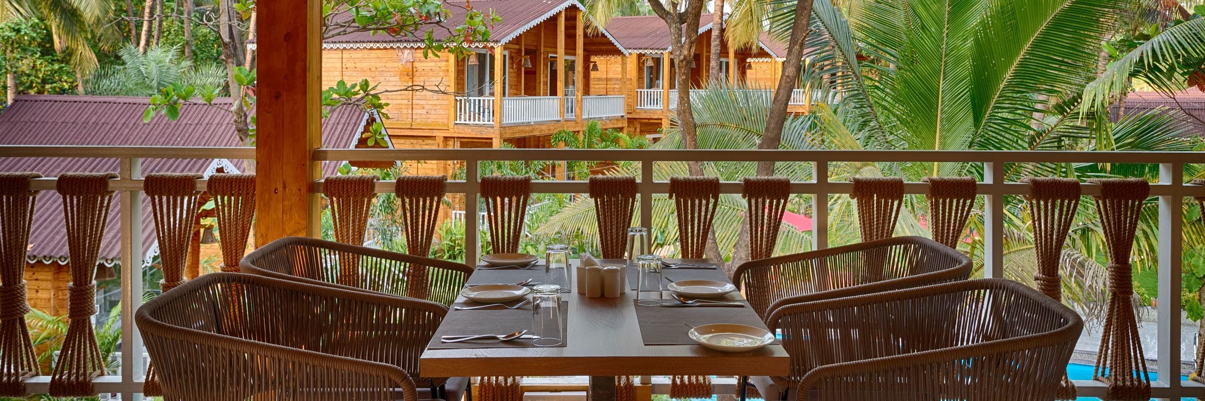 View from a covered dining area with woven chairs, a set dining table, overlooking wooden cabins among palm trees Aralea Beach Resort by Stone Wood, Morjim