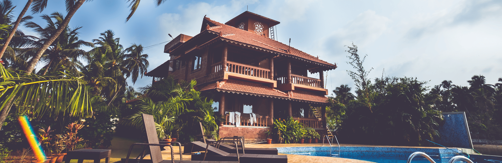 Red-roofed building with steps leading to a pool, surrounded by loungers & palm trees under blue sky at Paradise Lagoon Resort, Udupi.