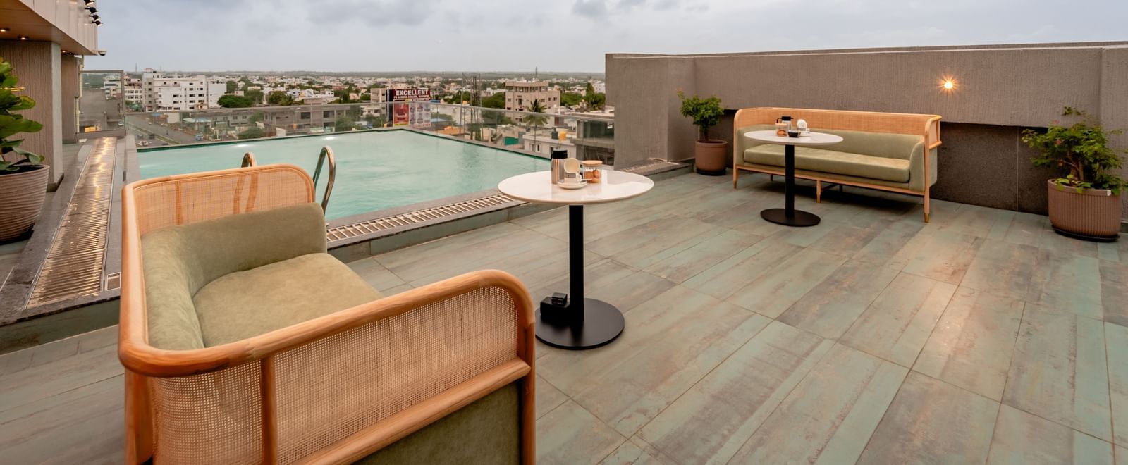 The rooftop swimming pool with clear blue water, surrounded by a tiled deck and some modern outdoor seating, offering a panoramic view of a city under a cloudy sky | Pravasa Hotel Vijayapura