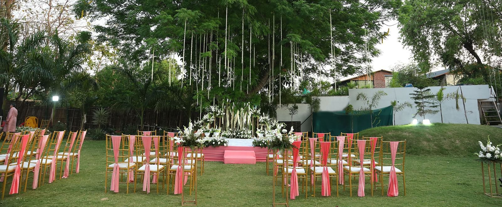 Daytime lawn view with red chairs and a large tree in the centre at Tisya Farms, Gurgaon.