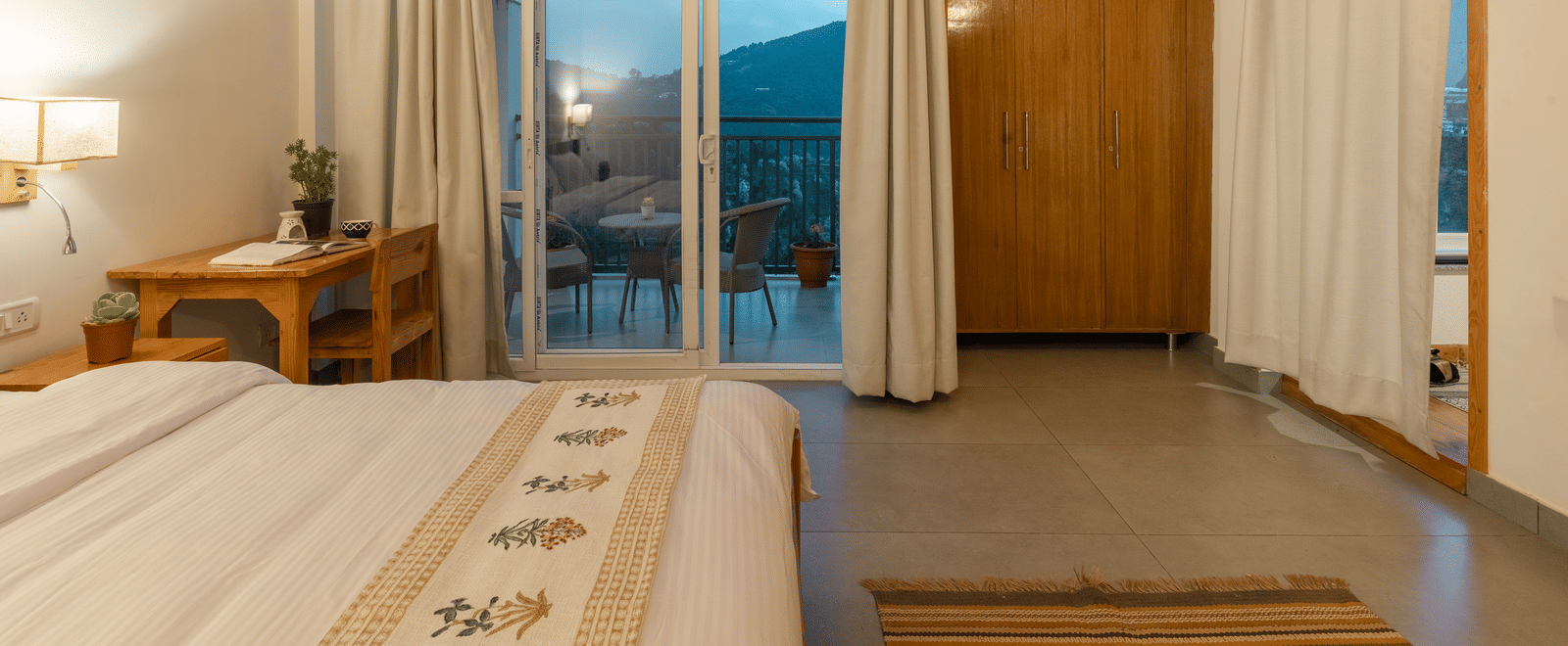 A close-up of a Ziran Retreat bedroom showing a bed with a white comforter, a bedside table with a lamp, and large glass doors leading to a balcony.