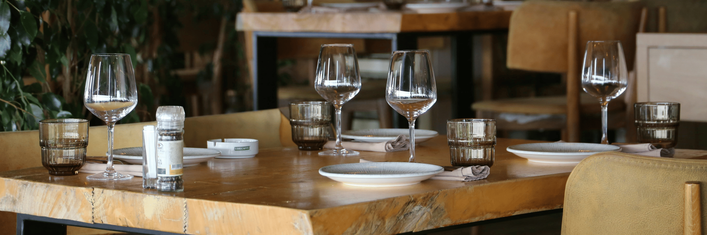 A thick wooden dining table set with wine glasses, plates, and napkins featuring green foliage in the background of the dimly lit restaurant.
