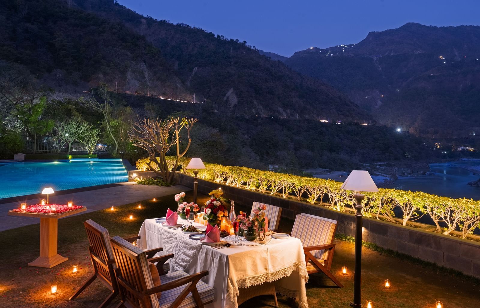 A dinner setup by a pool with mountains in the background. The table is adorned with candles and flowers.
