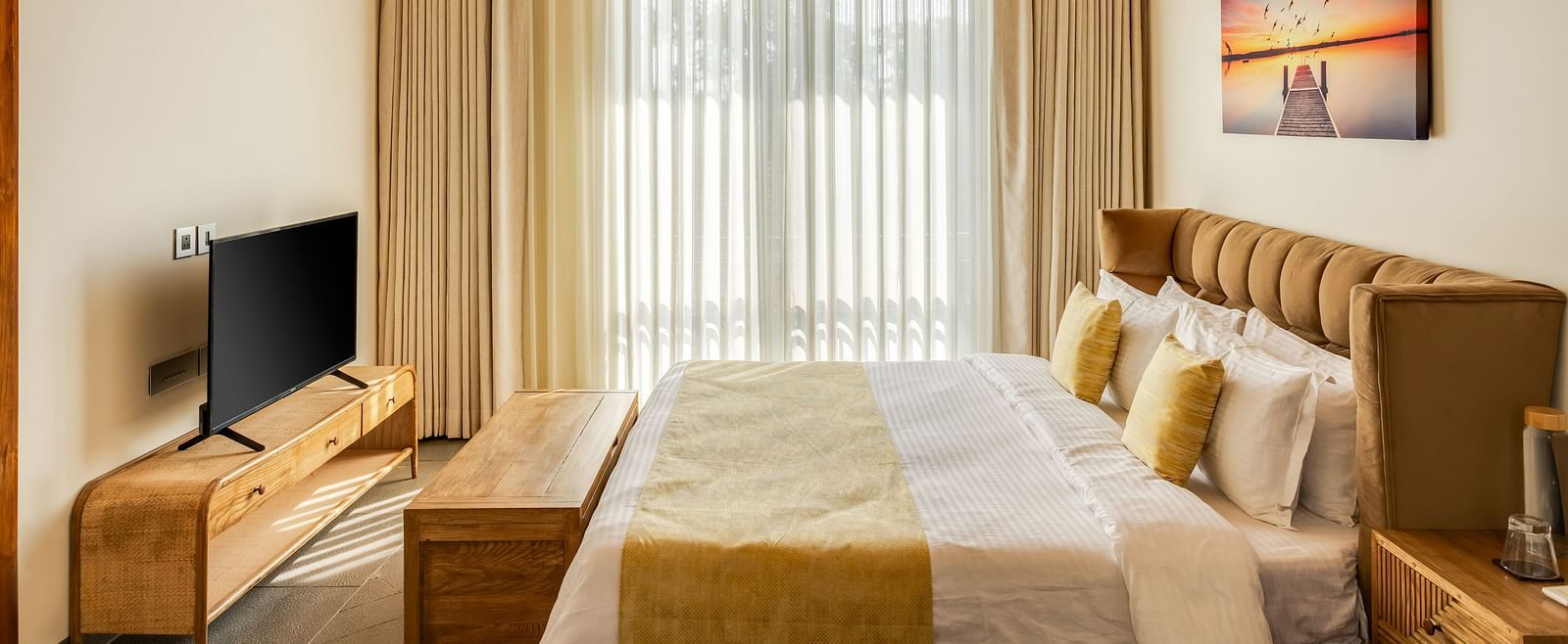 A cosy bedroom with twin beds, soft beige furnishings, a wall-mounted television, and a large window with flowing curtains.