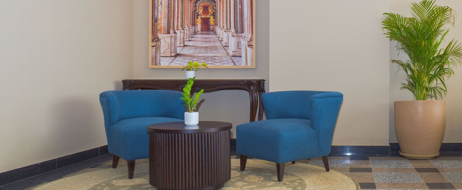 A pair of blue armchairs sit in a corner with a small table and a plant, beneath a painting of an archway - Urban Suites by BluSalzz, Jaipur
