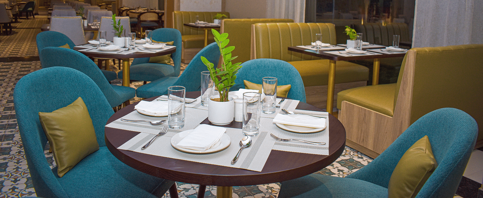 A restaurant interior with teal armchairs, booth seating, round tables, a patterned floor, sheer curtains, and hanging pendant lights - Urban Suites by BluSalzz, Jaipur