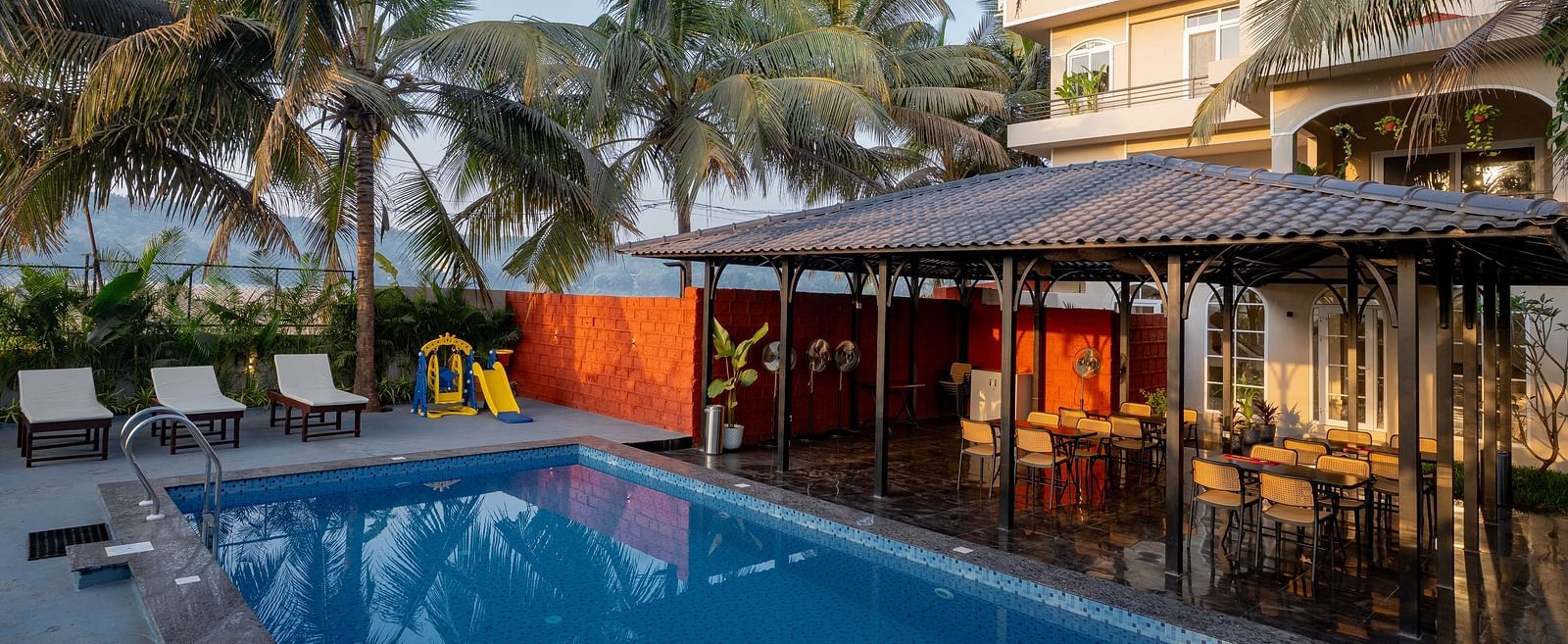 Outdoor swimming pool surrounded by palm trees, with a poolside seating area and Kssemya building in the background.