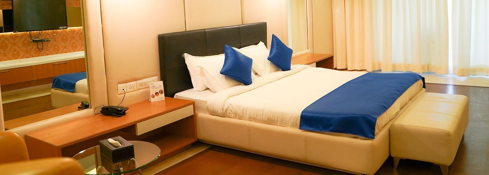 A warm-toned hotel room at BNGV Grandeur Hotel and Banquets, with a bed featuring blue and white linens, and soft lighting creating a cosy ambience.