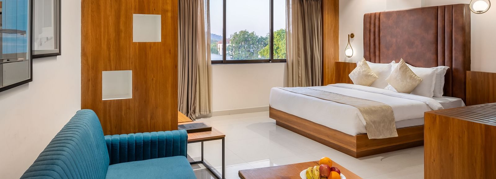 A well-lit hotel suite features a comfortable bed, a sofa, a wooden coffee table with fruit, and a window with curtains - Lords Eco Inn Ambaji.