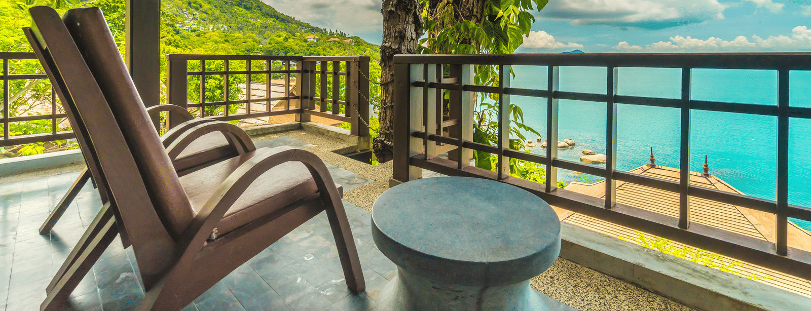 A balcony with 2 wooden lounge chairs, a small round table and a view of the sea and surrounding greenery.