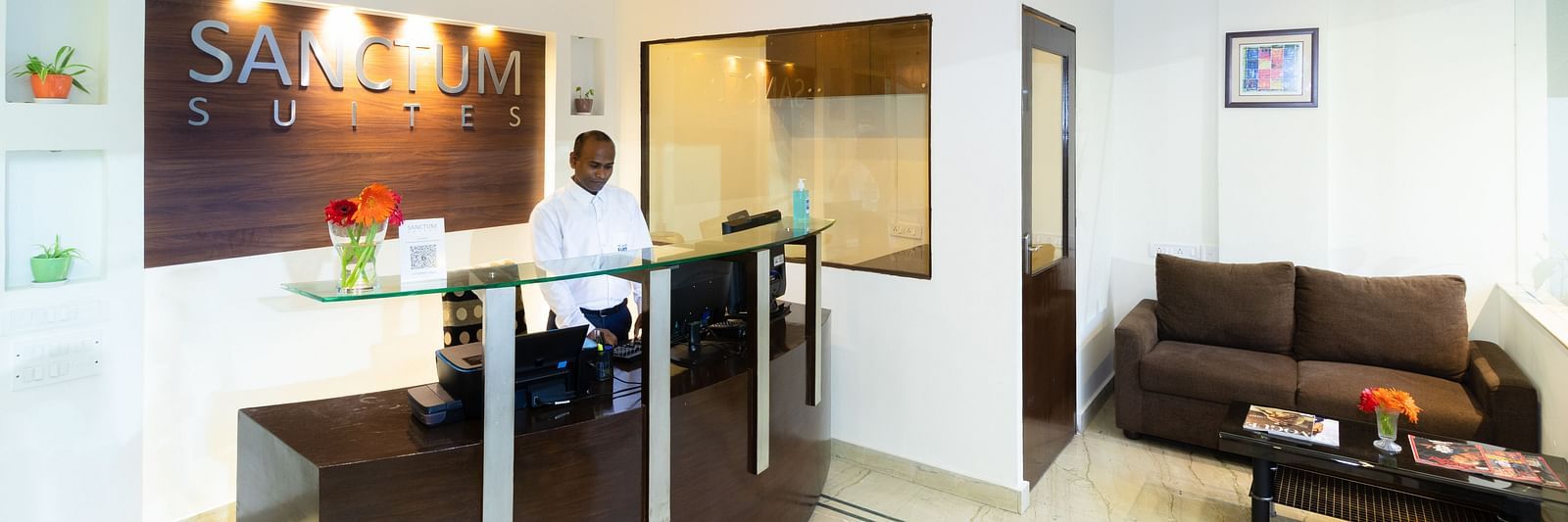 The reception area at Sanctum Suites Domlur, Bangalore featuring a reception desk, a brown sofa, and a coffee table.