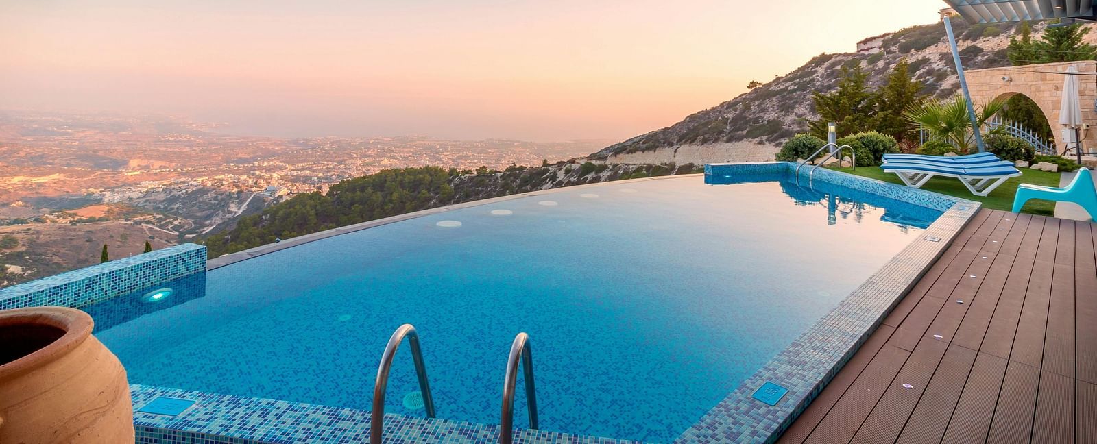 Infinity pool on a hillside with scenic views of the city and mountains at sunset, beside a wooden deck.