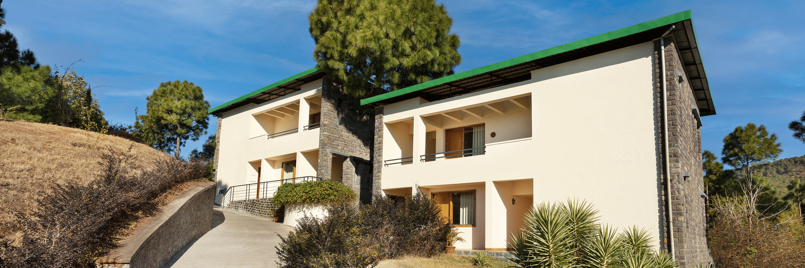 Exterior of premium rooms at Suryavilas Luxury Resort and Spa in Solan featuring the facade of the property on the mountain surrounded by trees and shrubs under a blue sky on a sunny day