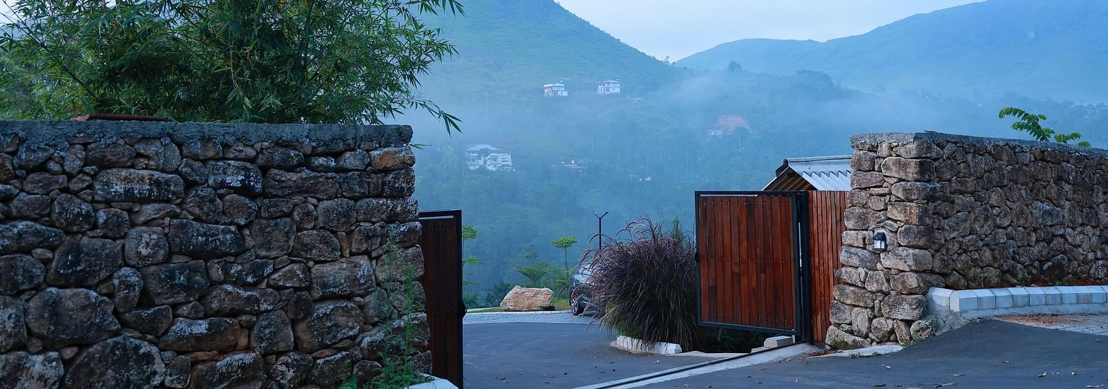 A stone wall entrance with an open wooden gate at Vaasta Vagamon, revealing a winding road and distant mist-shrouded hills.