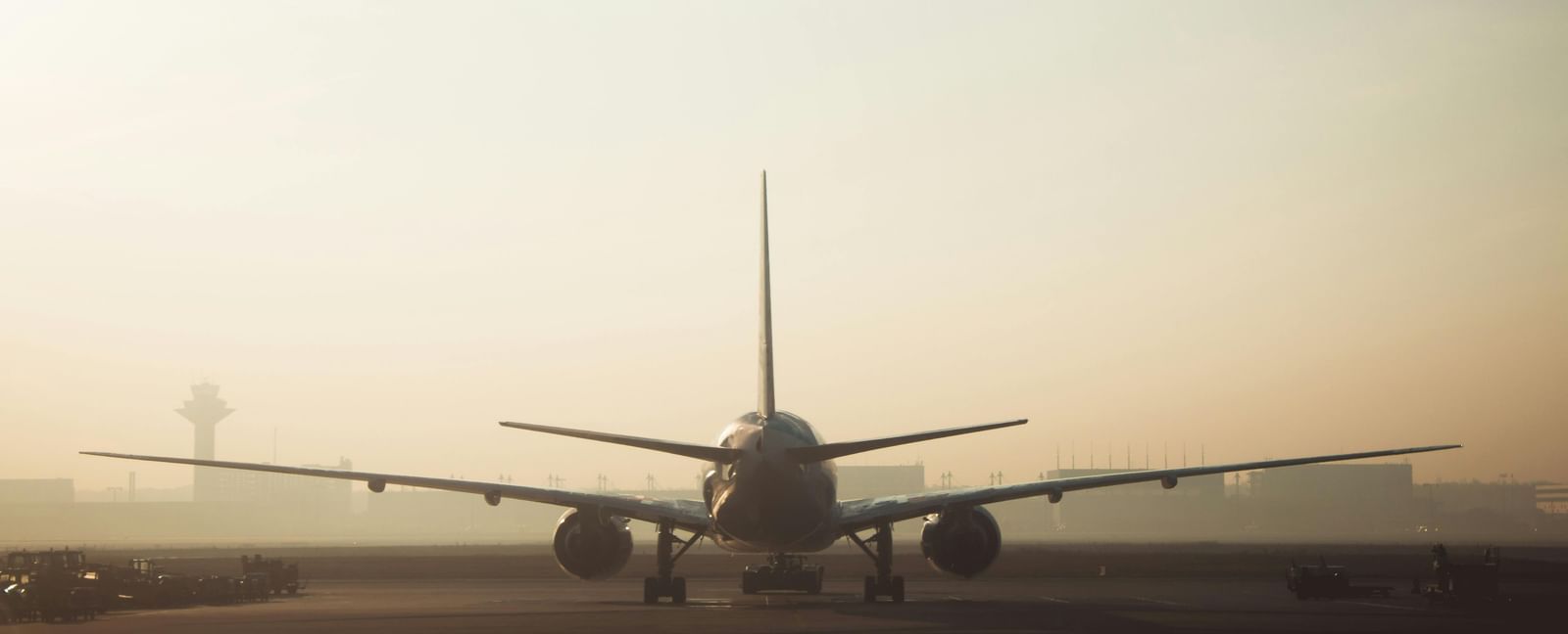 The rear view of a plane silhouetted against a hazy, light-filled sky at an airport, with the plane's tail fin and wings clearly visible.