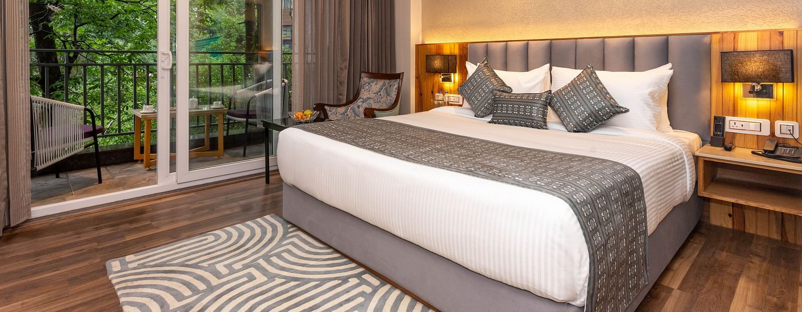 An image of a Superior Room with bed, bed side table, lamps, rug, and an expansive window leading to the balcony - The Belvedere Himalayan Retreat, McLeodganj