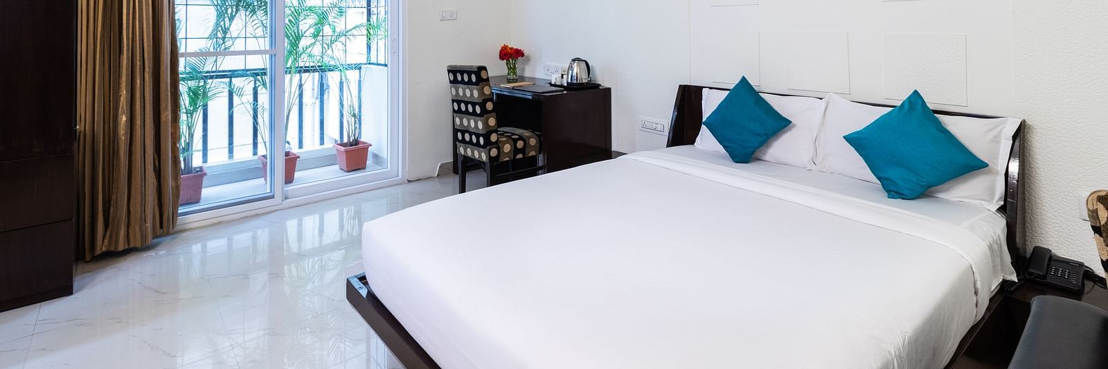 A room with a double bed with blue pillows, a work desk and chair, and glass door leading to the balcony at Sanctum Suites Domlur, Bangalore.