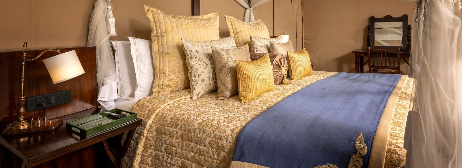 A large bed with pillows covered in golden coloured sheets and a lamp on a night stand by the side - Aalia Jungle Resort & Spa