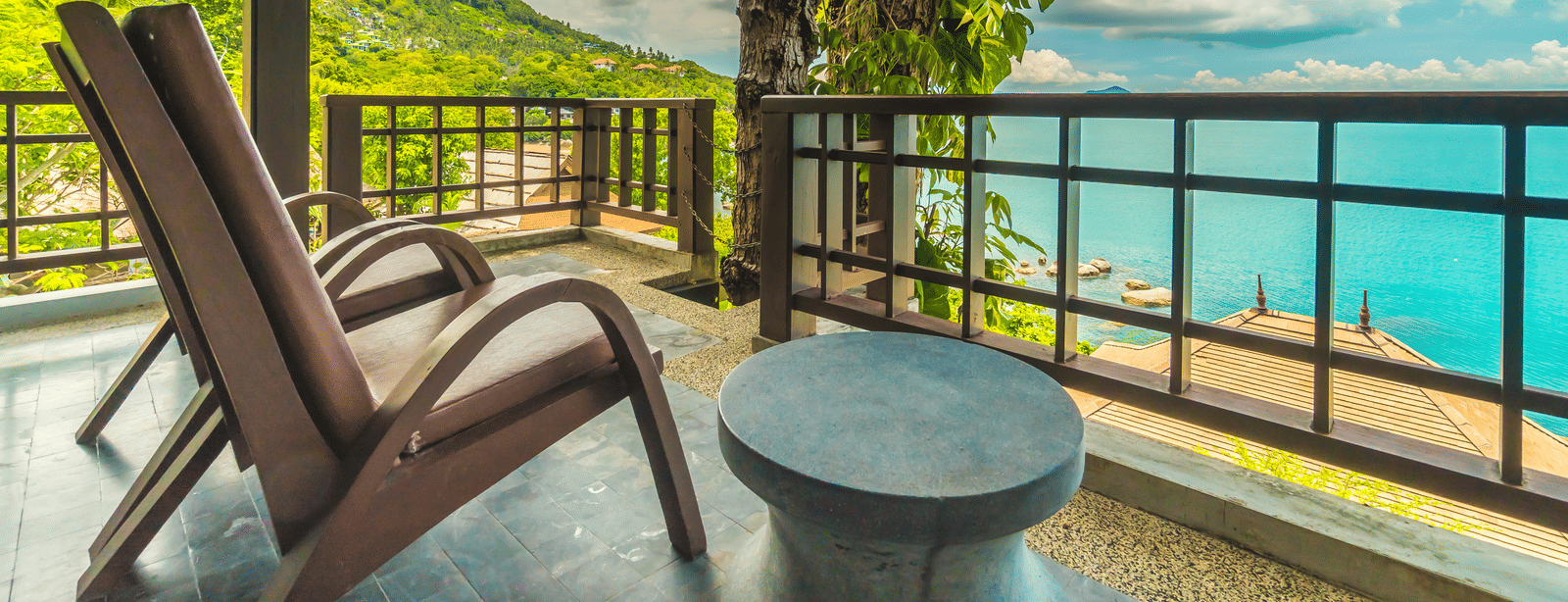 A balcony with 2 wooden lounge chairs, a small round table and a view of the sea and surrounding greenery.
