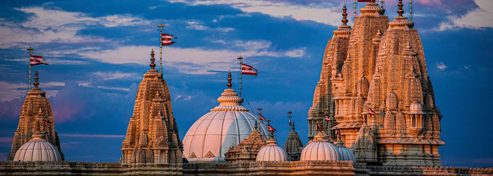 Close-up of temple spires (_shikharas_) at sunset, glowing warm orange against a dramatic, blurred blue and purple sky with small flags atop.