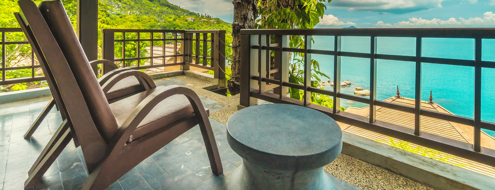 A balcony with 2 wooden lounge chairs, a small round table and a view of the sea and surrounding greenery.