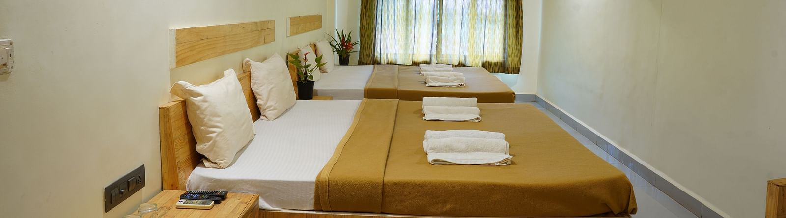 A side view of a room at Riverside Inn, featuring two beds placed beside each other, a bedside table, windows with curtains at the end, and a wall-mounted AC.