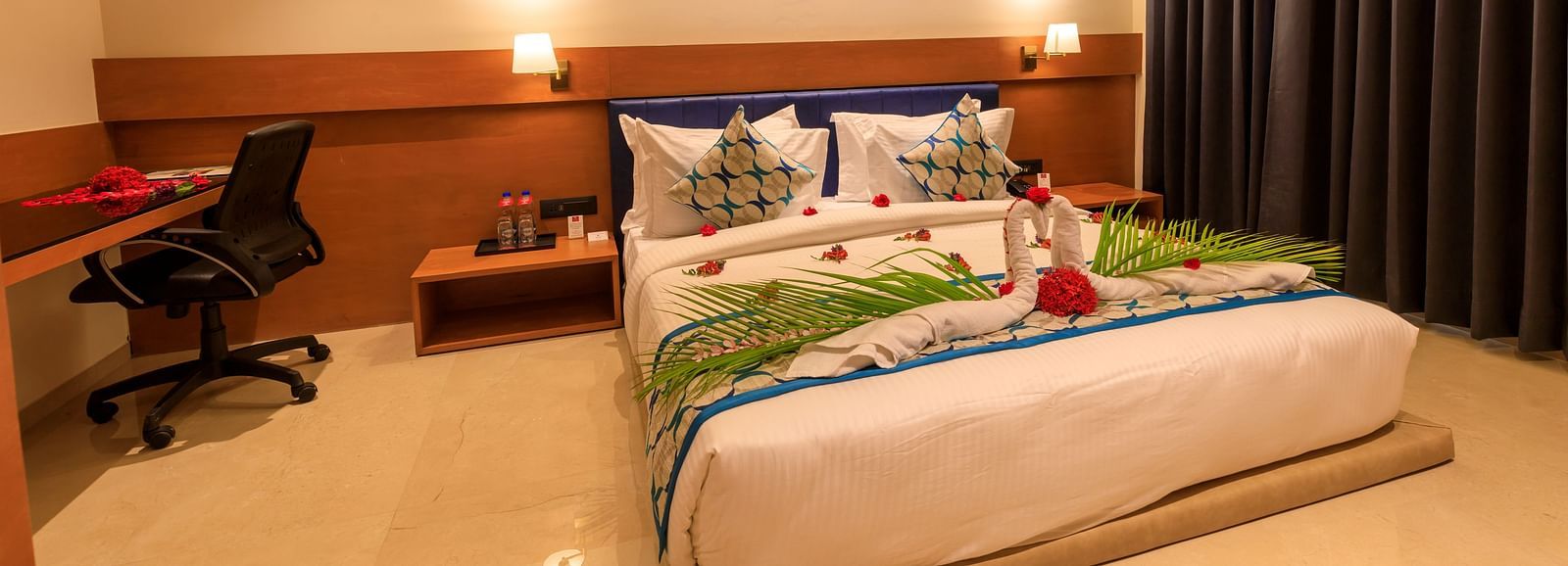 A warmly lit hotel room at Lords Eco Inn Apple Gate features a decorated double bed, bedside tables, a desk with a chair, and dark curtains.