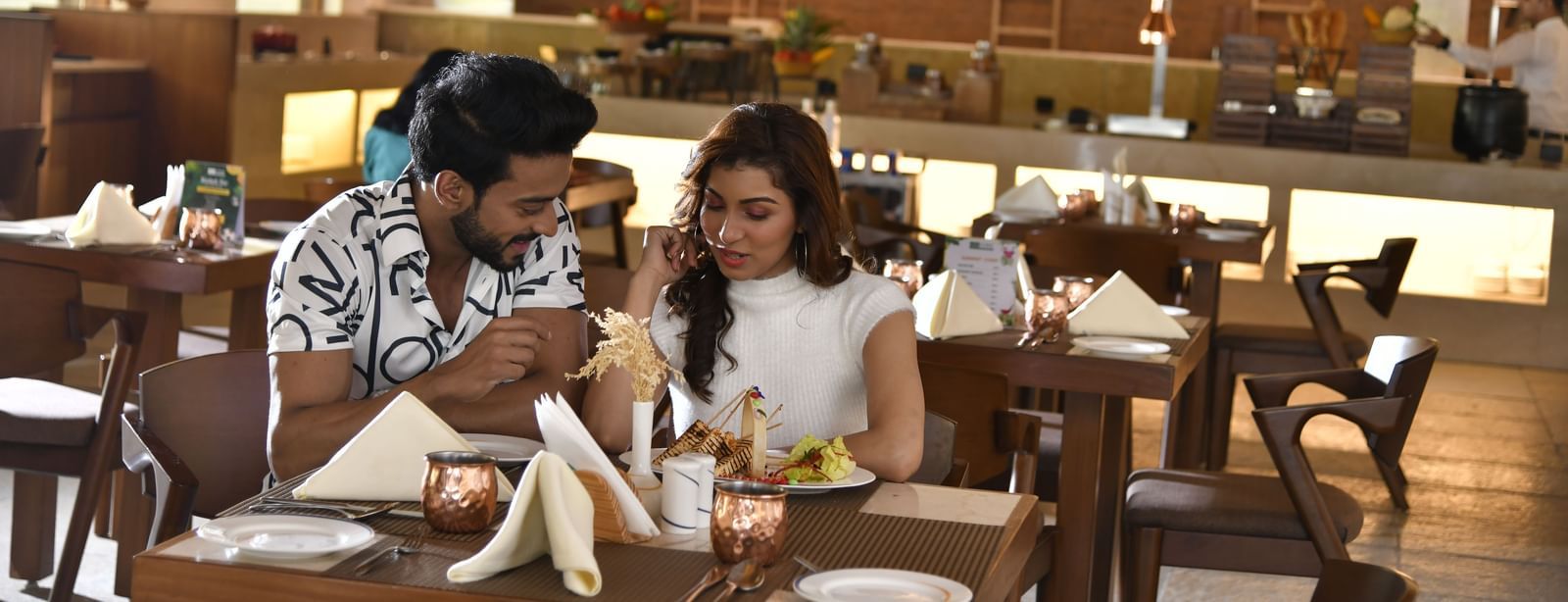 Two people are seated at a table in a dining area, engaged in conversation | Yagna