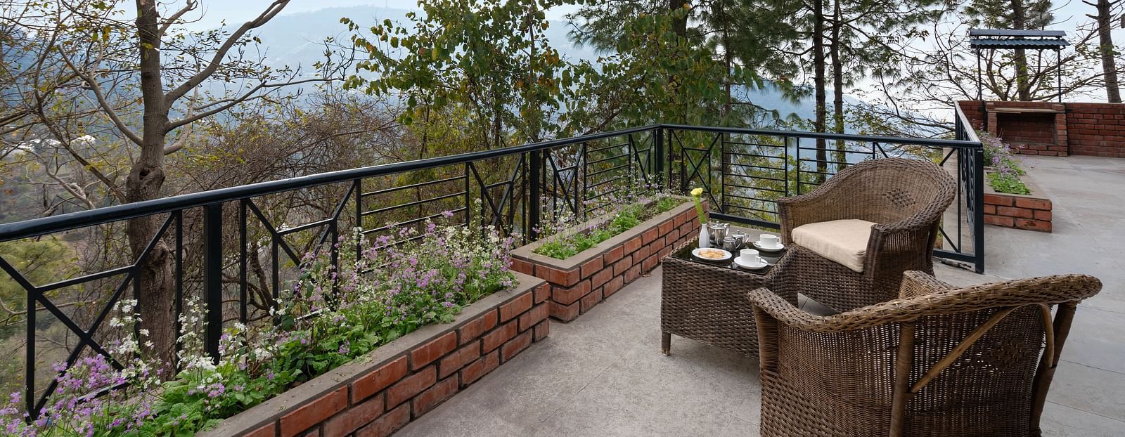Two wooden chairs and a table set on an open balcony with a view of trees and mountains afar - Baikunth resort, kasauli 