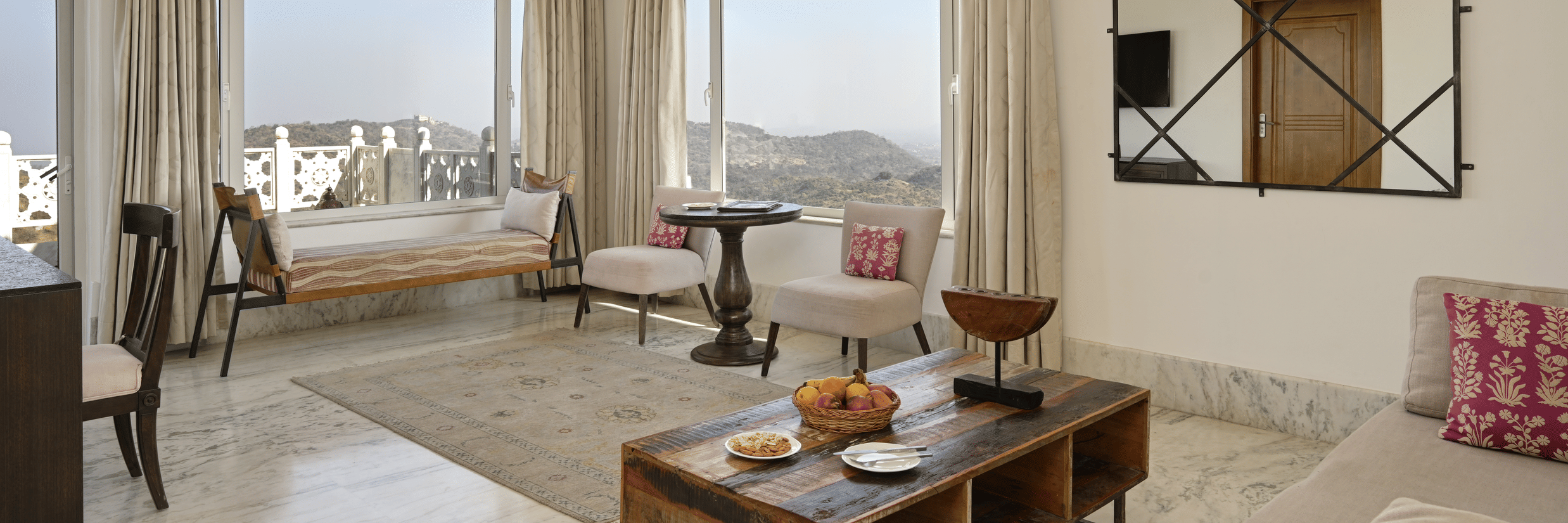 Plunge Pool Suite at Fateh Safari Suites featuring a seating area with sofa chairs, centre tables, work desk and chair, mirror on the wall, and windows which provides views of green hills.