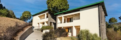Exterior of premium rooms at Suryavilas Luxury Resort and Spa in Solan featuring the facade of the property on the mountain surrounded by trees and shrubs under a blue sky on a sunny day