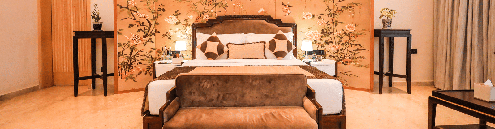 A room shows a bed with side tables, plants on both sides, a bench at the foot of the bed and a wall panel behind the bed at Ananta Spa & Resort, Jaipur.