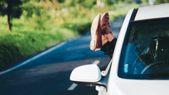 A person with their legs out of the window of a car parked on a road with trees in the background.