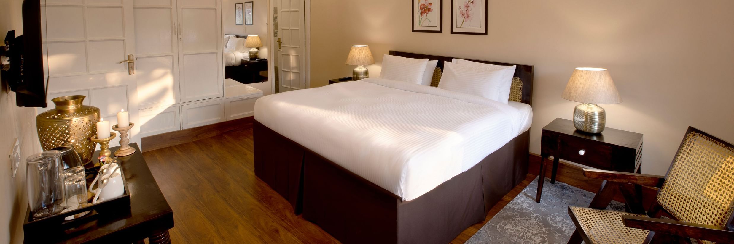 An image of super deluxe room featuring a king size bed, bedside table on either side, a flat TV screen, a table where antique items are placed, and  a chair in one corner of the room- Lamrin Boutique Cottages Rishikesh