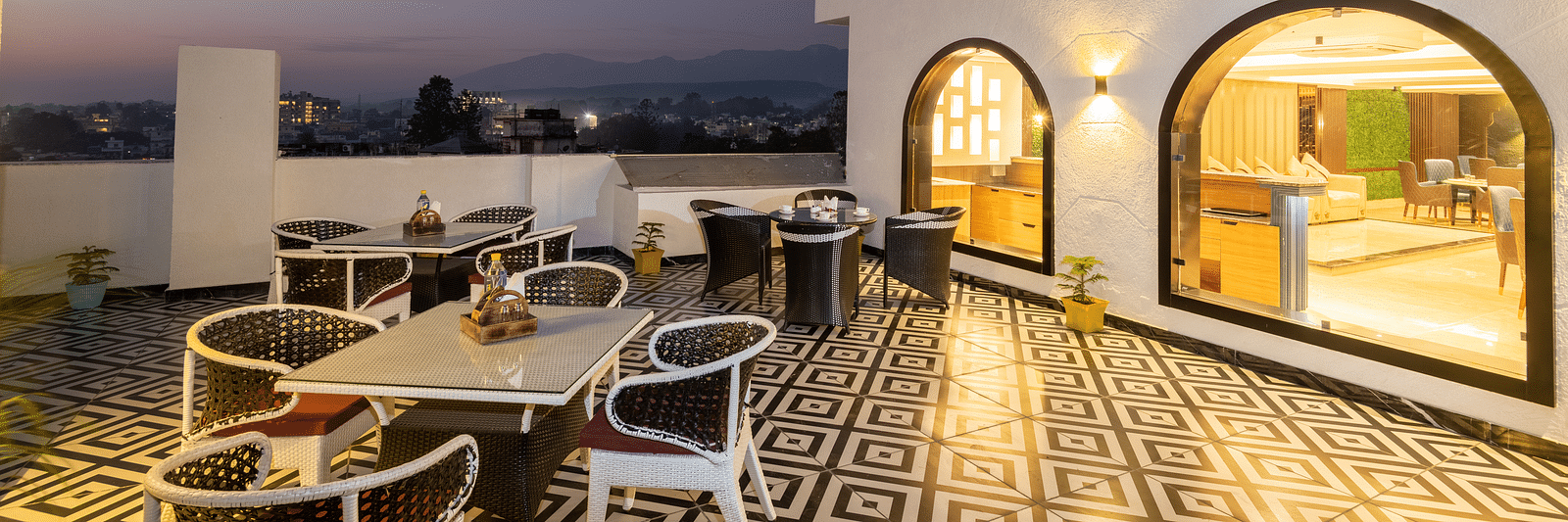 Image of the rooftop restaurant at DLS Grand Luxotica, Dehradun during evening featuring chairs and tables arranged on a well-furnished floor