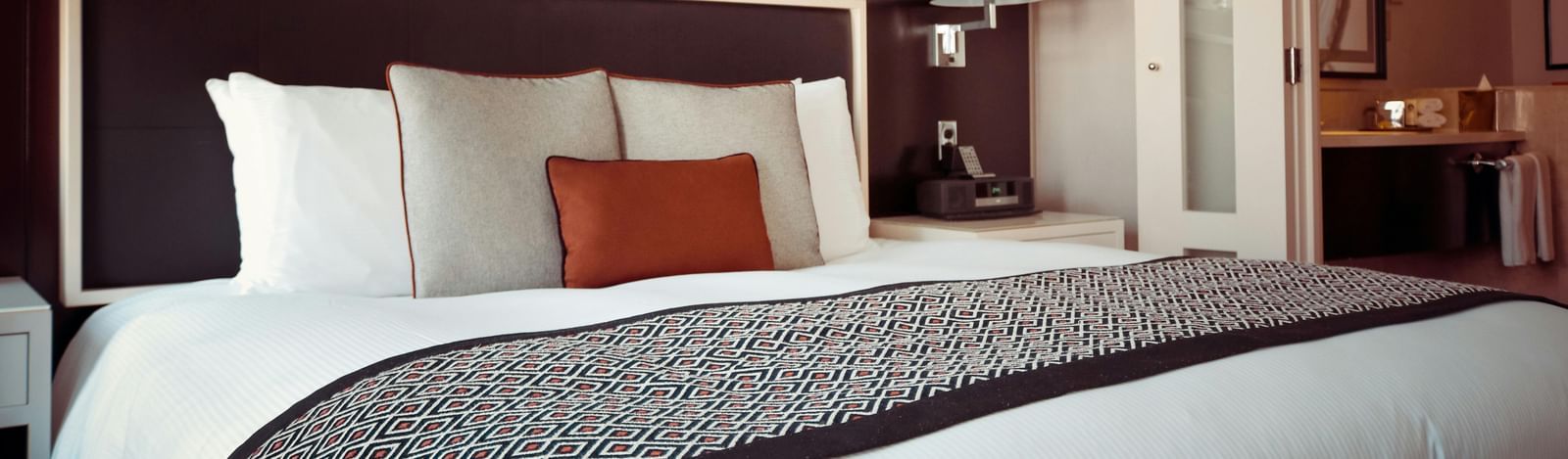 A hotel room displays a dark headboard, white linens, and a pop of colour with an orange pillow.