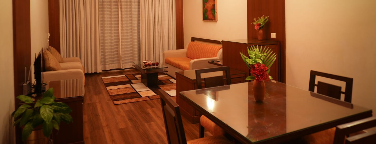 Living area of the One-Bedroom Suite at Vedic Village Spa Resort, showcasing plush sofas, wooden flooring, closed drapes, and a well-lit dining space.