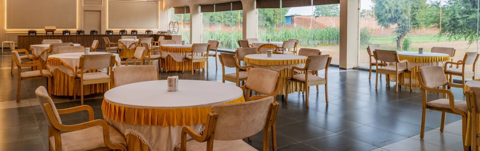 Spacious banquet area with large windows and round tables surrounded by chairs at Hotel Highway King, Neemrana. 