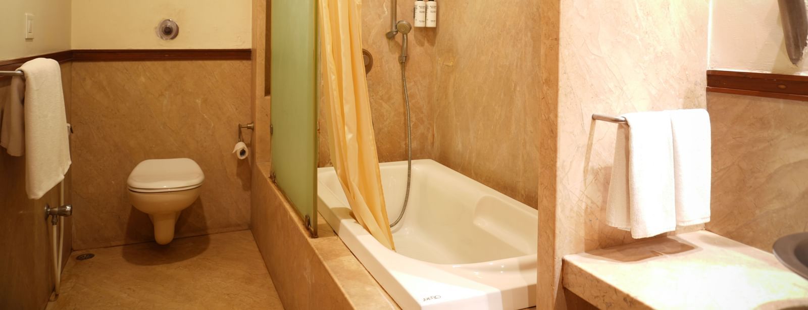 Washroom in the One-Bedroom Suite at Vedic Village Spa Resort with wall-mounted western commode, bathtub, sink, and towels neatly placed on railings.