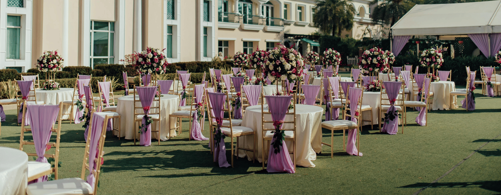 An outdoor event setup with rows of tables and chairs decorated on a lawn in front of a large building.