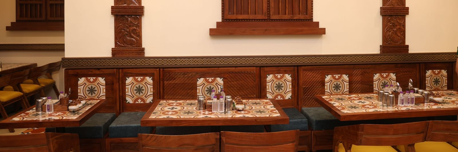 Indoor dining setup with traditional wooden décor, intricately carved pillars, tiled table settings, and cushioned wooden chairs with yellow seats in a warmly lit restaurant.