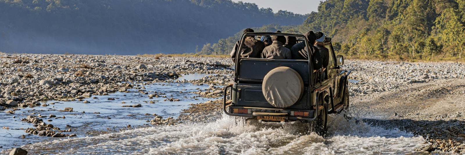 A jeep driving through a river in Jim Corbett National Park with forested hills in the background.