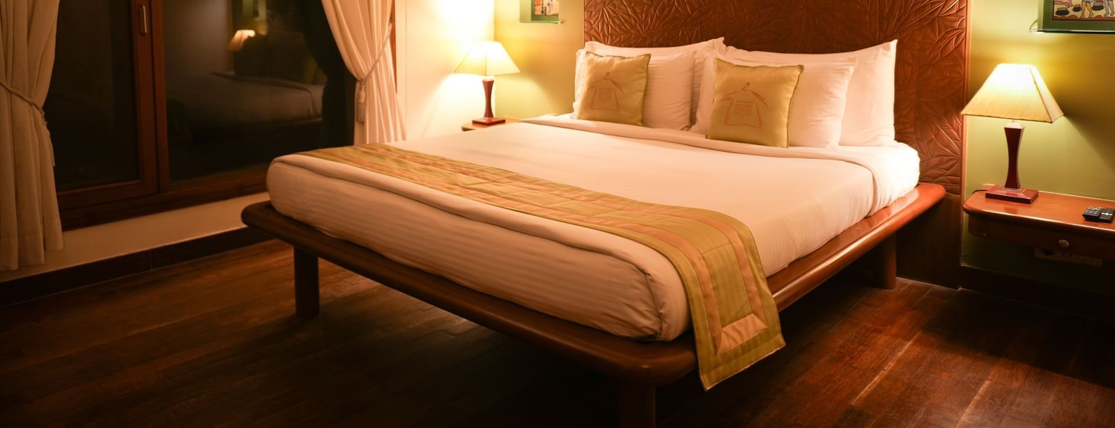 Interiors of the Lake View Villa at Vedic Village Spa Resort featuring a queen size bed, large windows with rolled-up blinds, warm lighting, wooden flooring, and framed paintings on the wall.