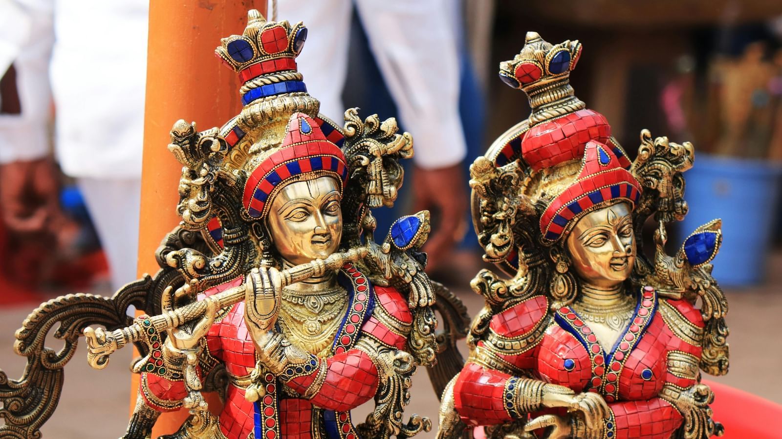 Two intricate brass statues of the Hindu deities Radha and Krishna, adorned with red and blue details. Krishna is playing a flute.