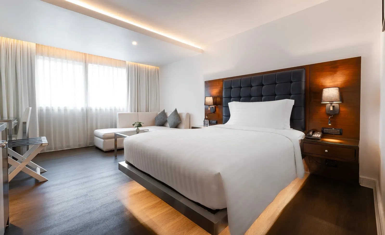 Modern hotel bedroom with tufted navy headboard, platform bed, white seating area, walnut accents, and floor-to-ceiling curtains at Night Hotel Bangkok.