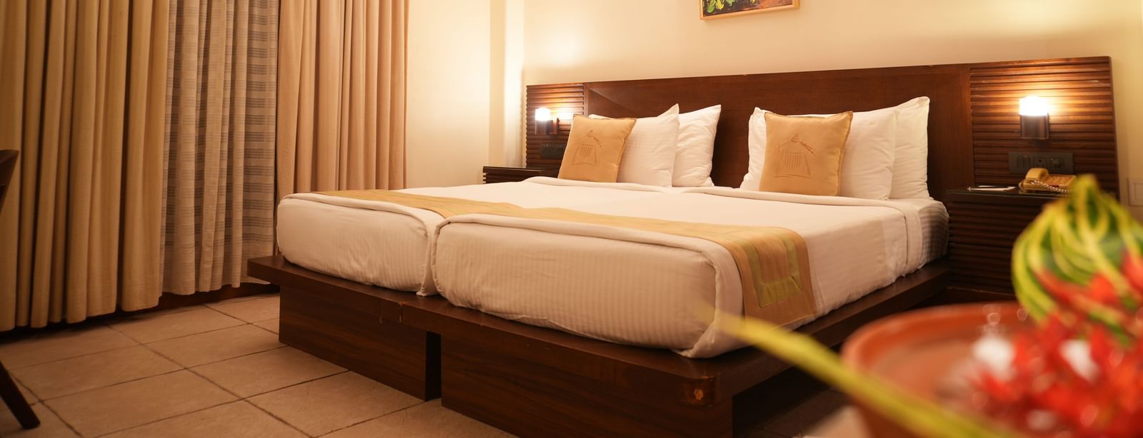 A warmly lit Superior Room at Vedic Village Spa Resort featuring a double bed with a wooden headrest, drapes over a large window, and a painting above the bed.