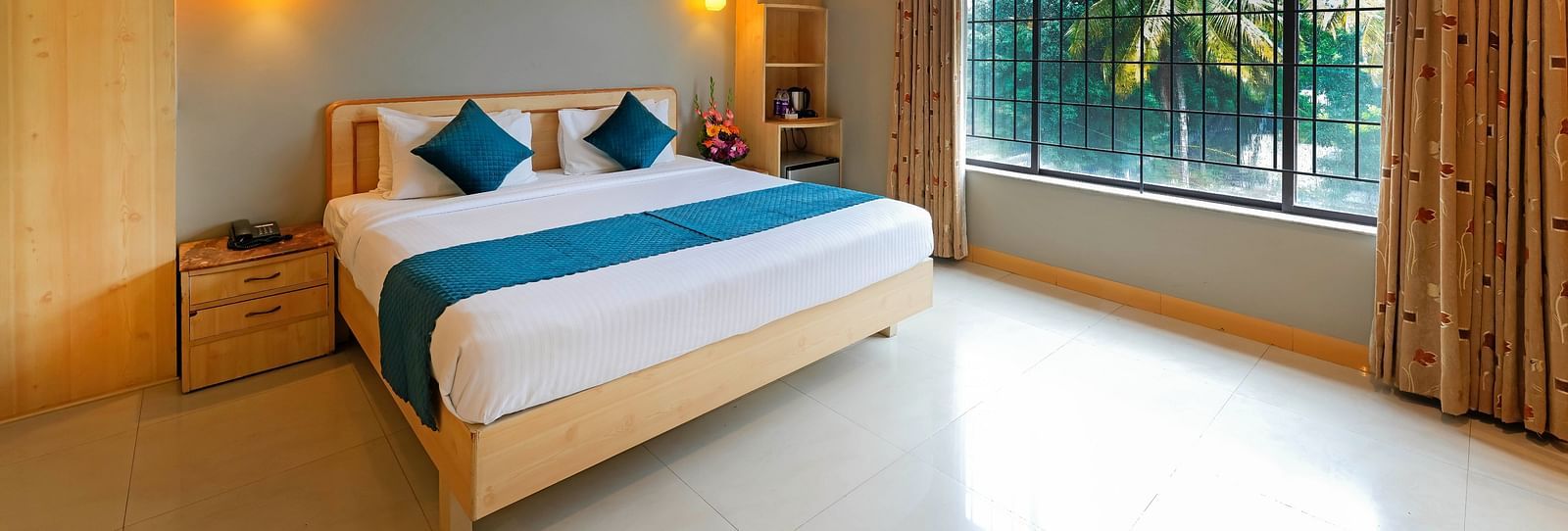 An image of a hotel room in Bengaluru featuring a queen size bed, a night stand, and sunlight from the expansive windows - Cross Roads Inn, Bengaluru