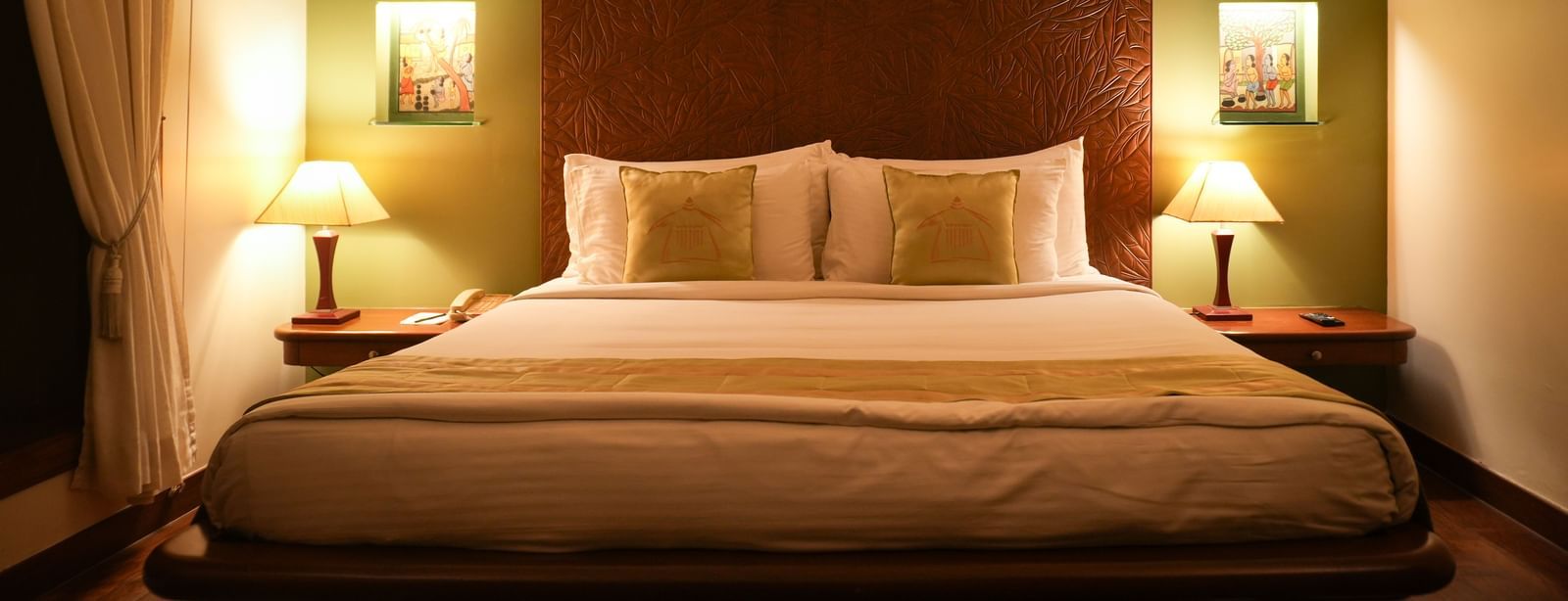 Front angle view of the bedroom in the Lake View Villa at Vedic Village Spa Resort showing a neatly made queen size bed and a framed painting under soft lighting.