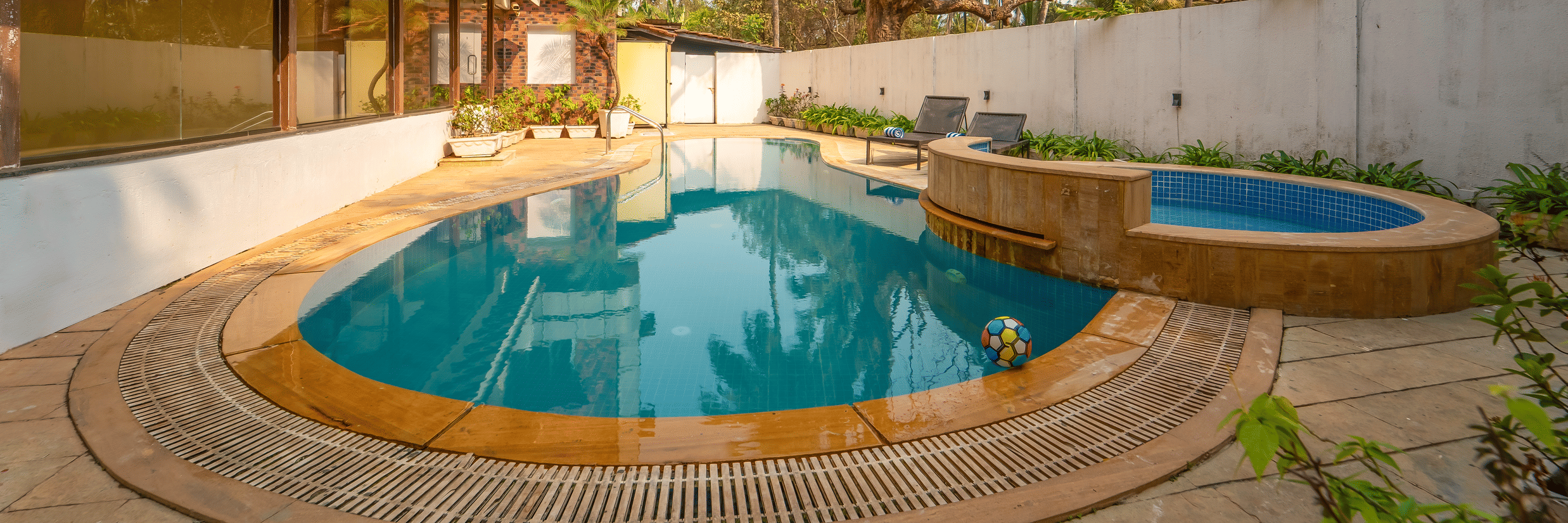 Rosetum Anjuna's kidney-shaped pool and raised circular jacuzzi in daylight, flanked by lounge chairs, a colourful beach ball in the water, and mature trees.